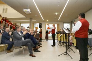 Musikaufführung in einem modernen Veranstaltungsraum, dirigiert von einem Mann in Tracht, mit applaudierendem Publikum und Kinderchor.