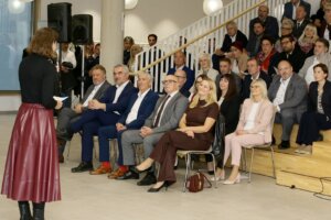 Person spricht vor einer Gruppe von Menschen auf einem Event; Zuhörer*innen sitzen aufmerksam in Reihen.