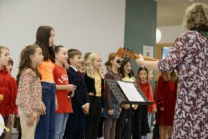 Kinderchor-Auftritt mit Gitarrenbegleitung in einer Schulaula, festgehalten für einen Artikel im regionalen Prima Magazin.