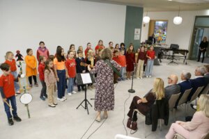 Eine Kindergruppe singt bei einer Schulaufführung, begleitet von einer Lehrerin mit Gitarre, während Eltern im Publikum sitzen.