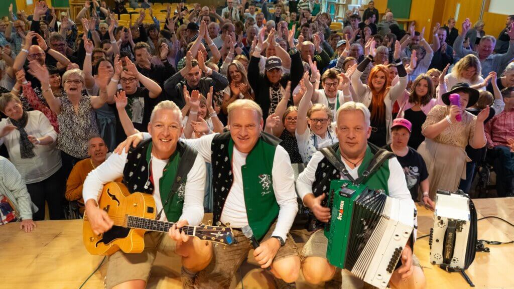 Musikgruppe in traditioneller Tracht spielt vor fröhlichem Publikum bei einem lokalen Event, strahlende Gesichter im Prima Magazin.
