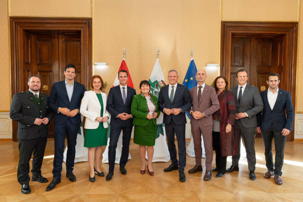 Gruppe von Männern und Frauen in formeller Kleidung vor Flaggen in einem eleganten Raum, Gruppenfoto für Prima Magazin.
