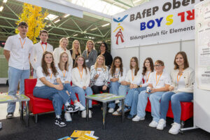 Gruppe von Schülern und Lehrern sitzt lächelnd auf einer Messe, repräsentiert die Bildungseinrichtung BAfEP Oberösterreich.