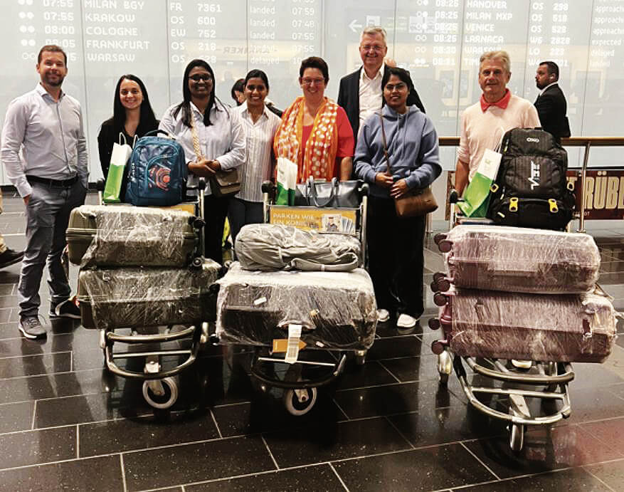 Gruppe von Reisenden am Flughafen mit Koffern und Taschen, lächelnd vor Ankunftstafel, ideal für Prima Magazin.