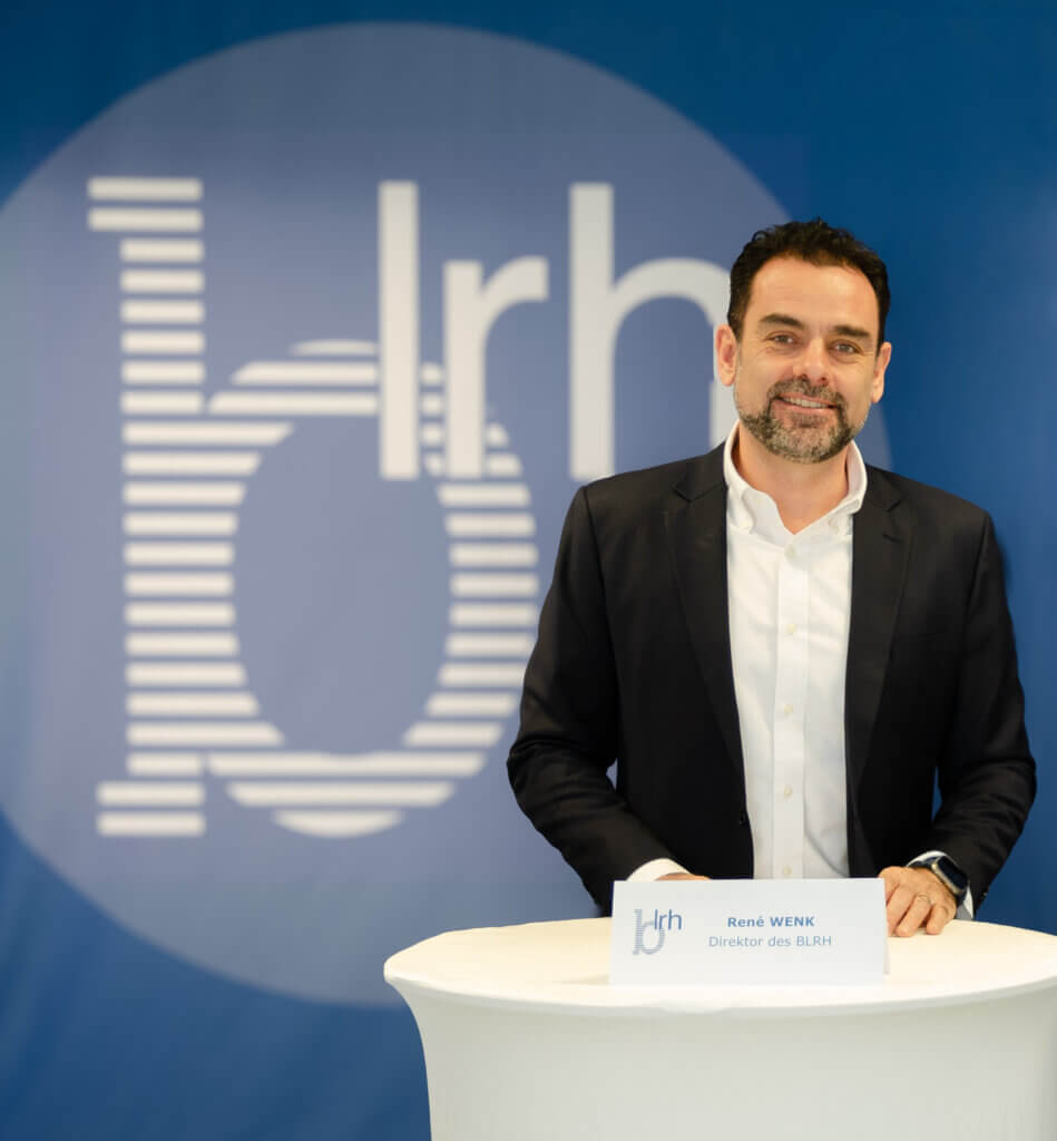 Ein Mann im Anzug steht lächelnd vor einem blauen BLRH-Hintergrund und einem Tisch mit Namensschild. Foto zum Thema Finanzschulden Burgenland