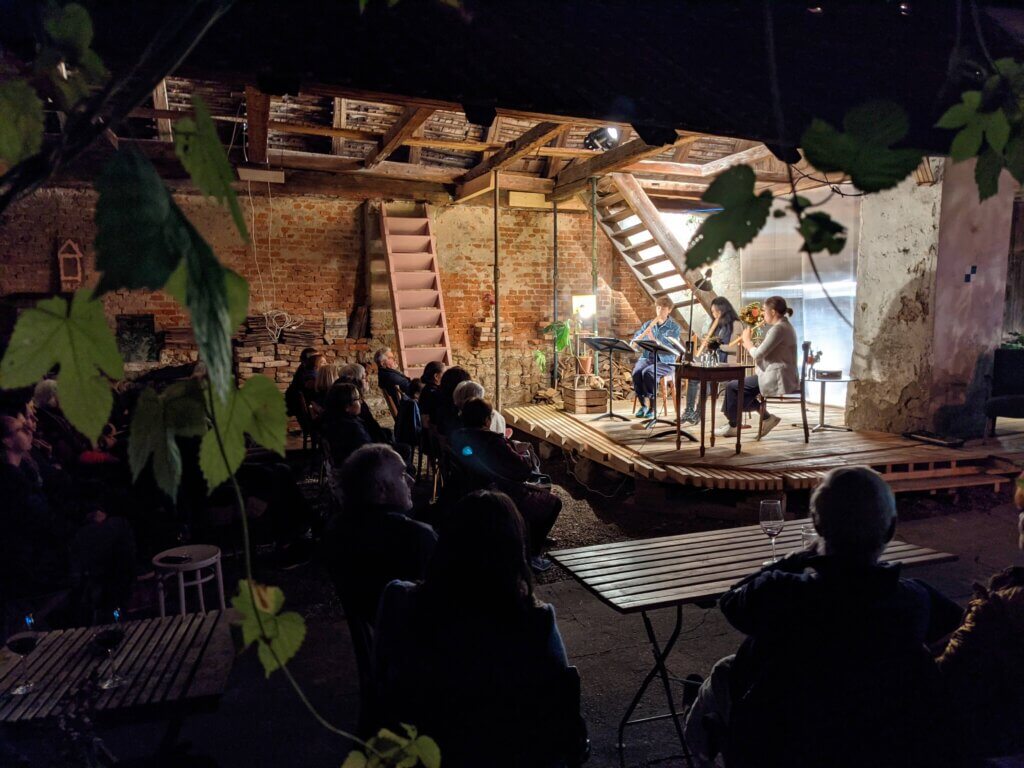 Konzert in gemütlicher Scheune, Musiker auf Bühne, Publikum sitzt an Holztischen, entspannte Atmosphäre, Prima Magazin Event.