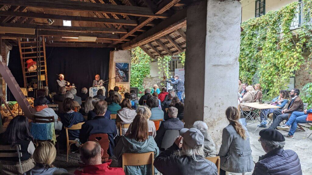 Lesung in rustikalem Innenhof. Publikum lauscht aufmerksam zwei Männern auf einer Bühne. Atmosphäre von Gemeinschaft und Kultur.