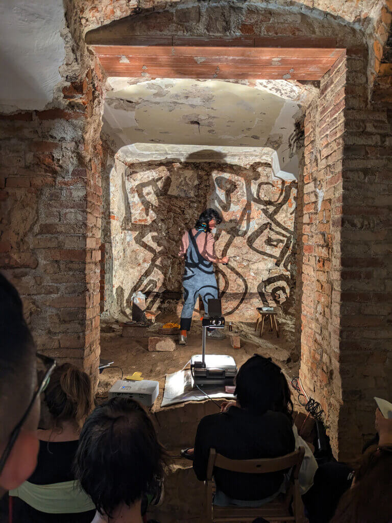 Künstler sprüht Graffiti in einem Gewölbekeller, während Zuschauer gespannt die kreative Performance im regionalen Magazinformat verfolgen.