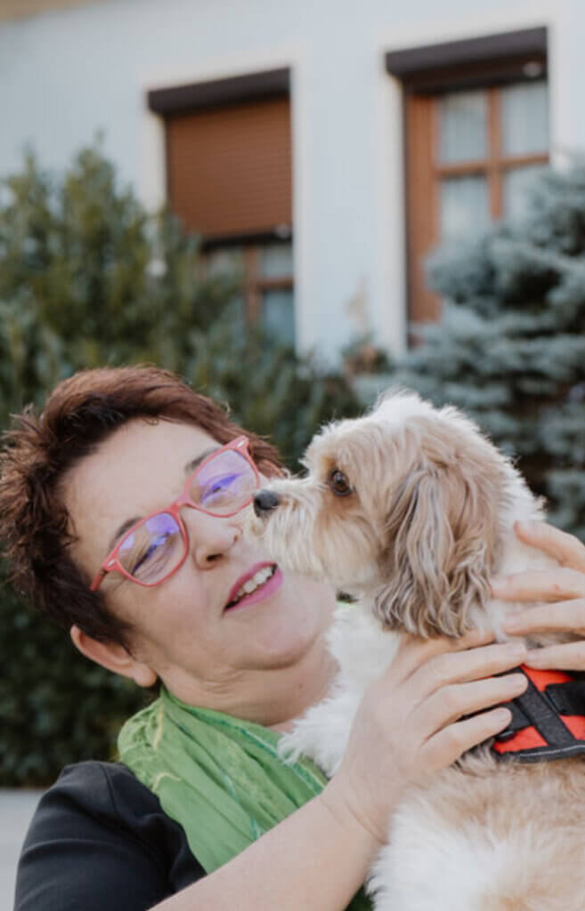 Ältere Frau mit roter Brille hält fröhlich einen kleinen Hund im Arm vor einem Haus im Freien.