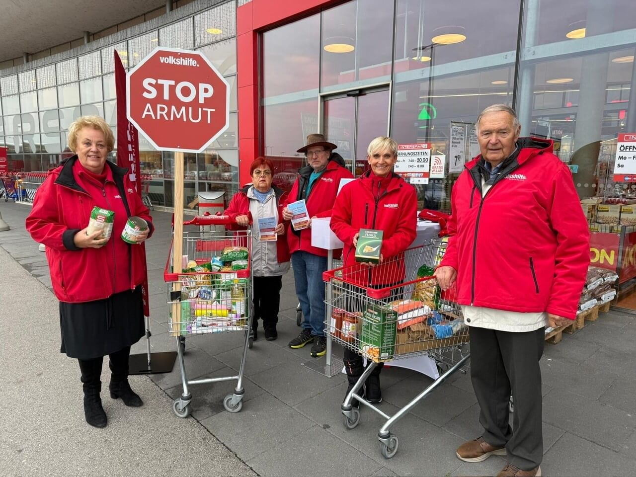 Gruppe von Freiwilligen in roten Jacken organisiert eine Lebensmittelspendeaktion vor einem Geschäft, Stop Armut-Schild im Hintergrund.
