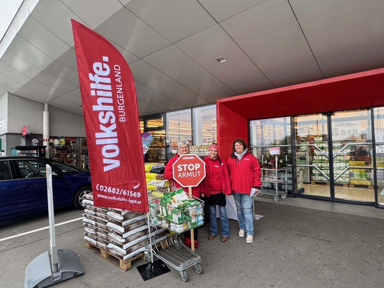 Personen in roten Jacken sammeln vor einem Supermarkt Spenden für Volkshilfe Burgenland. Banner daneben informiert über Armut.