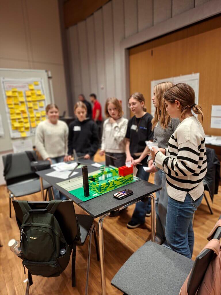 Gruppe von Jugendlichen diskutiert über ein LEGO-Modell bei einem Workshop in einem Konferenzraum, geeignet für Prima Magazin.