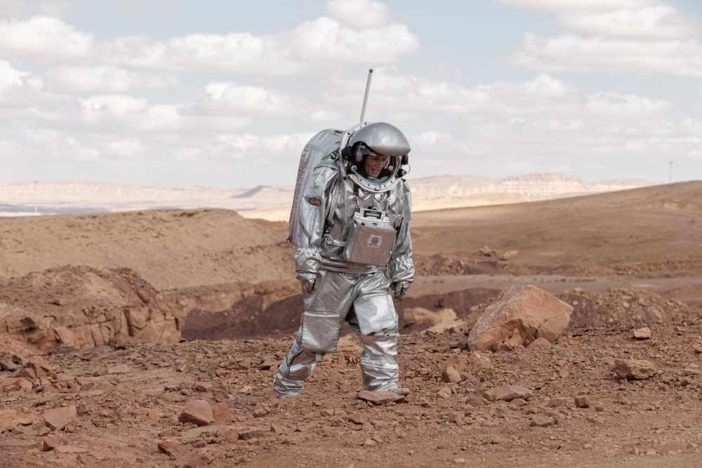 Astronaut in silbernem Anzug auf felsiger, marsähnlicher Landschaft unter blauem Himmel mit Wolken.