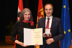 Person erhält Auszeichnung mit Urkunde und Medaille vor Flaggenhintergrund. Feierliche Ehrung im regionalen Kontext.