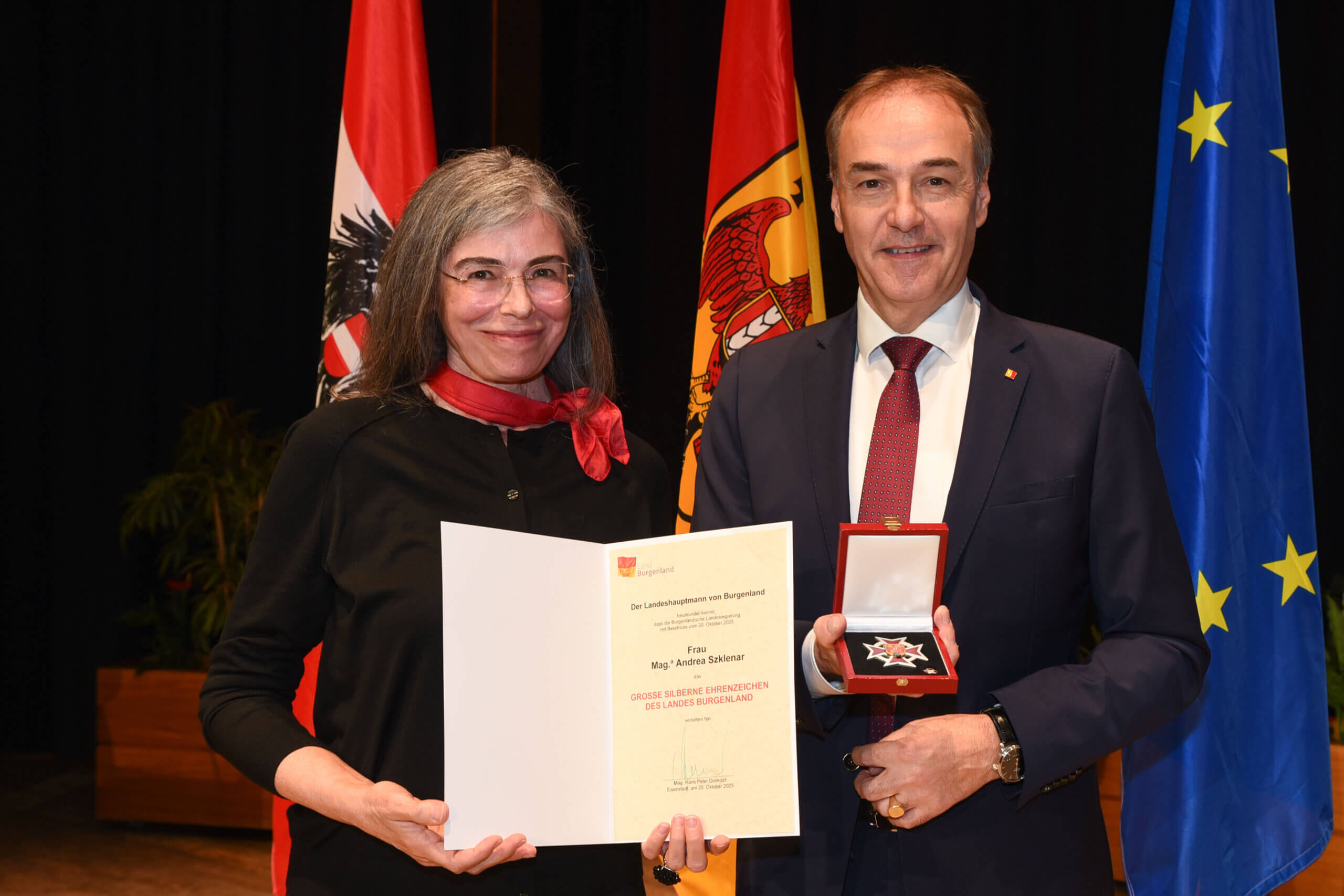 Person erhält Auszeichnung mit Urkunde und Medaille vor Flaggenhintergrund. Feierliche Ehrung im regionalen Kontext.