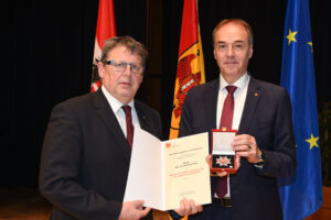 Zwei Männer im Anzug bei einer Auszeichnung mit österreichischen und EU-Flaggen im Hintergrund, einer hält ein Zertifikat.