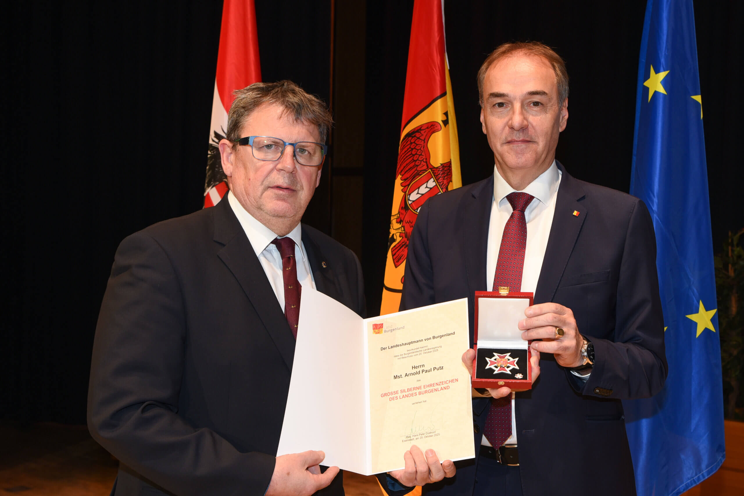 Zwei Männer im Anzug bei einer Auszeichnung mit österreichischen und EU-Flaggen im Hintergrund, einer hält ein Zertifikat.