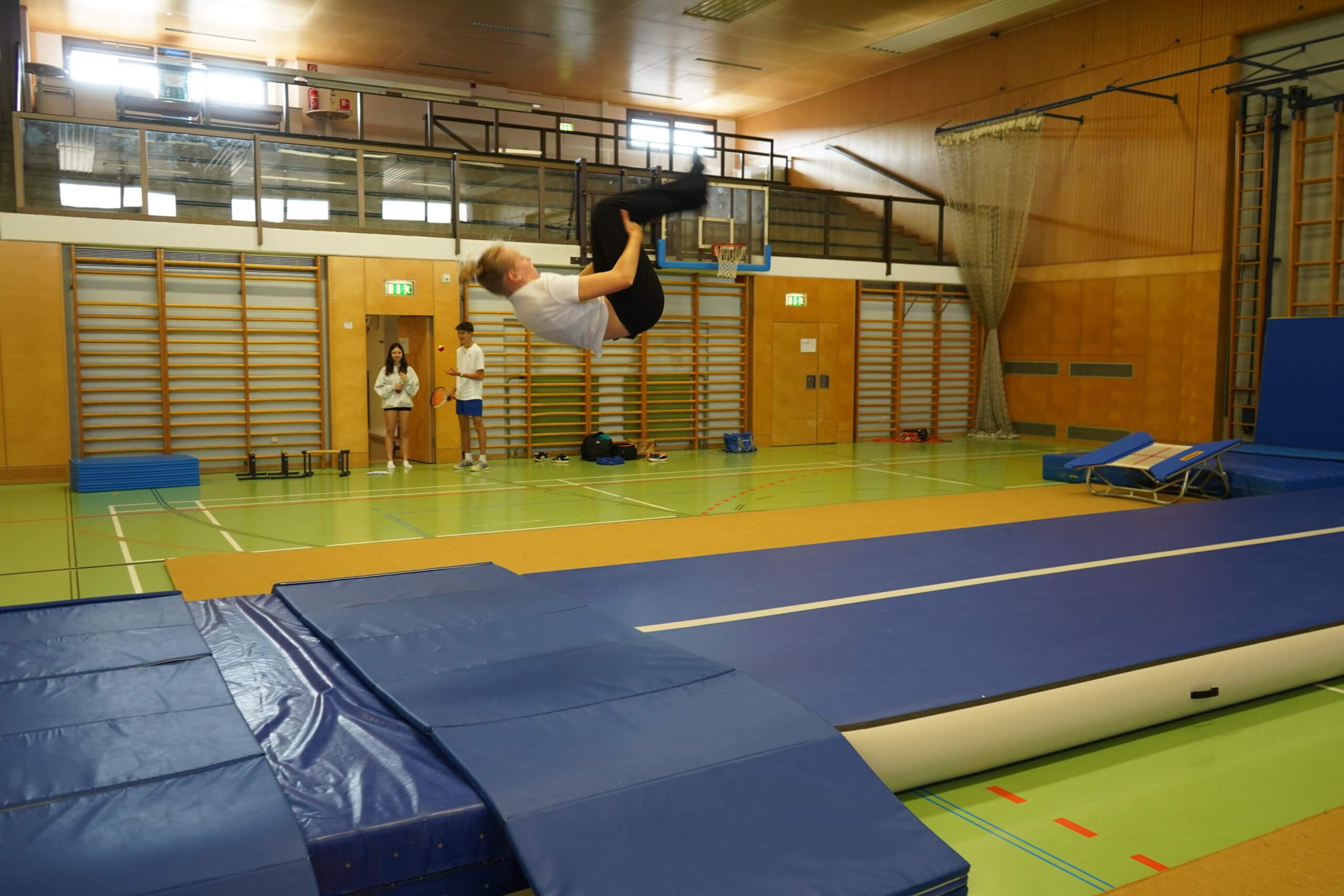 Ein Schüler zeigt einen Rückwärtssalto in der Turnhalle, während zwei andere zusehen, sportliche Atmosphäre im Prima Magazin.