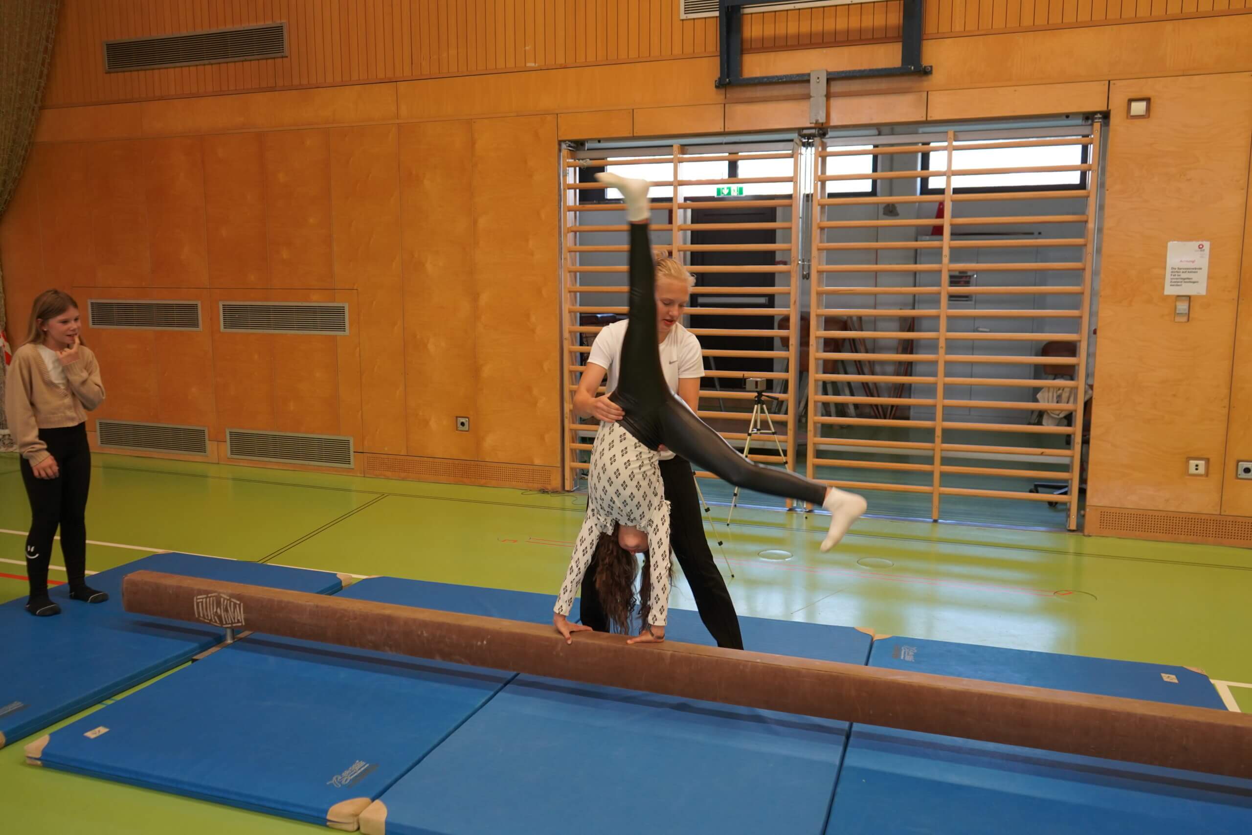 Mädchen übt in Turnhalle Handstand auf Schwebebalken, betreut von Trainerin, Zuschauerin schaut zu.