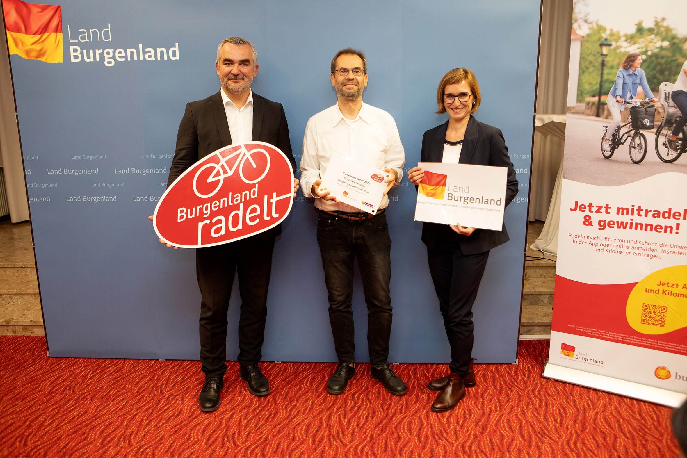 Drei Personen präsentieren Burgenland radelt-Schilder vor einem blauen Hintergrund und einem Werbebanner.