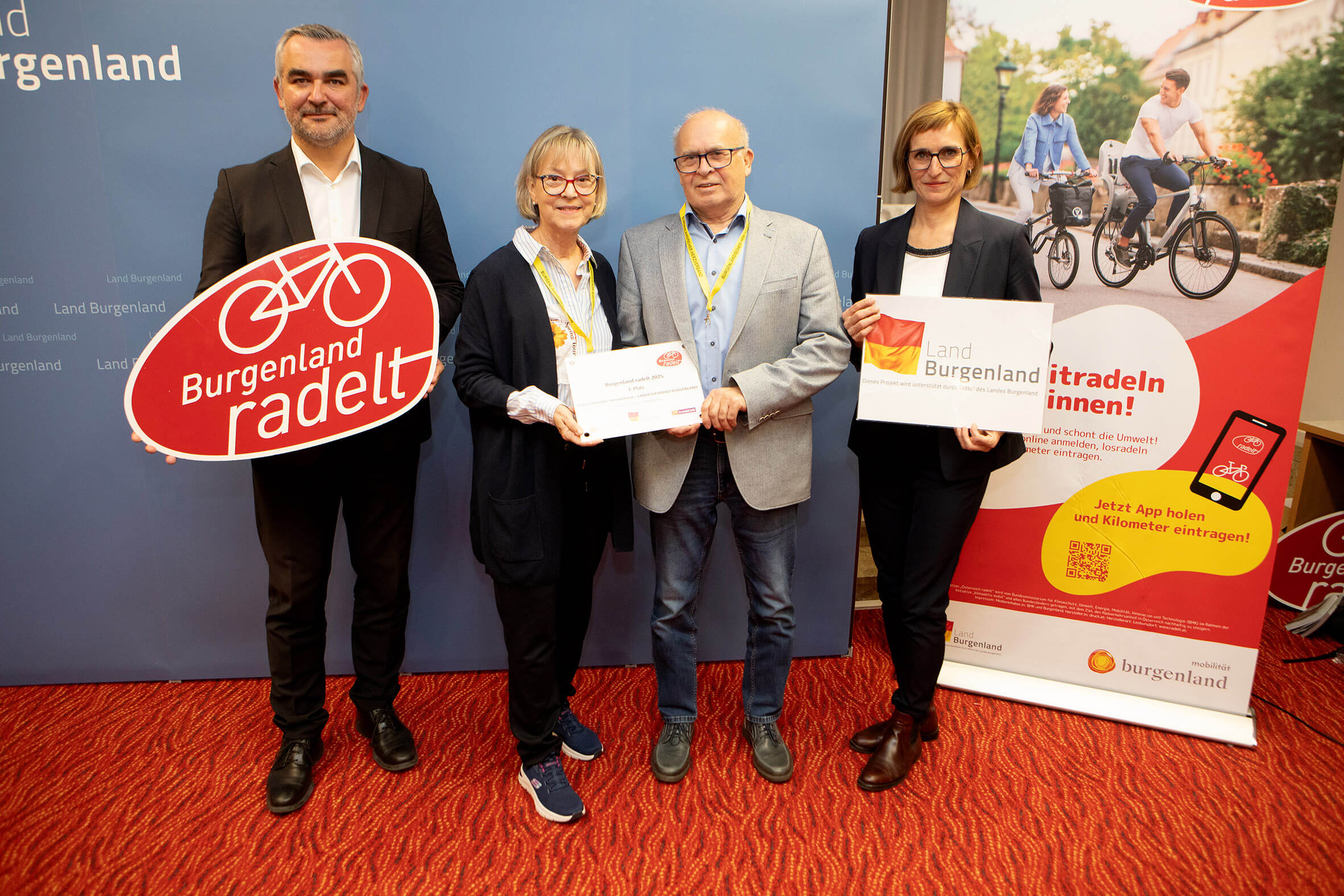 Vier Personen präsentieren das Burgenland radelt Projekt mit Schildern, die zur Teilnahme an der Radfahrkampagne aufrufen.