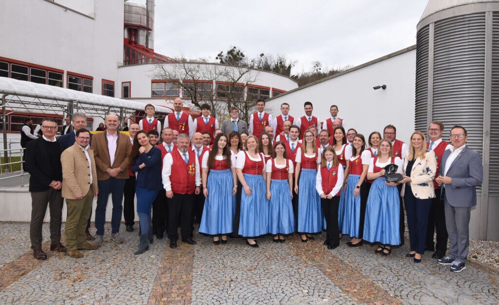 Eine Trachtengruppe posiert fröhlich vor einem modernen Gebäude. Regionale Gemeinschaft und Tradition im Fokus.