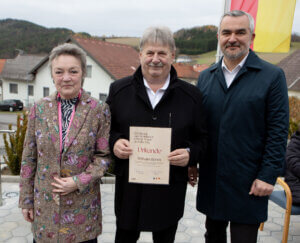 Drei Personen bei einer Zeremonie im Freien, eine hält eine Urkunde. Hintergrund mit Gebäuden und Landschaft.