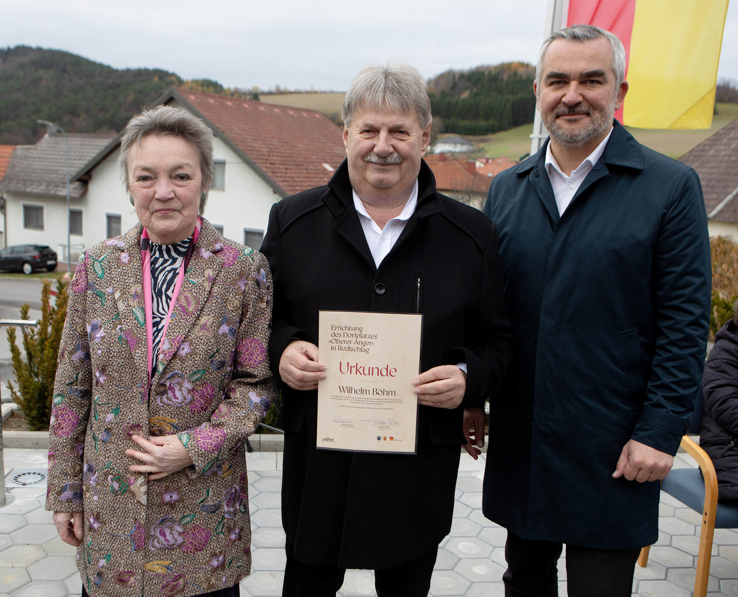 Drei Personen bei einer Zeremonie im Freien, eine hält eine Urkunde. Hintergrund mit Gebäuden und Landschaft.