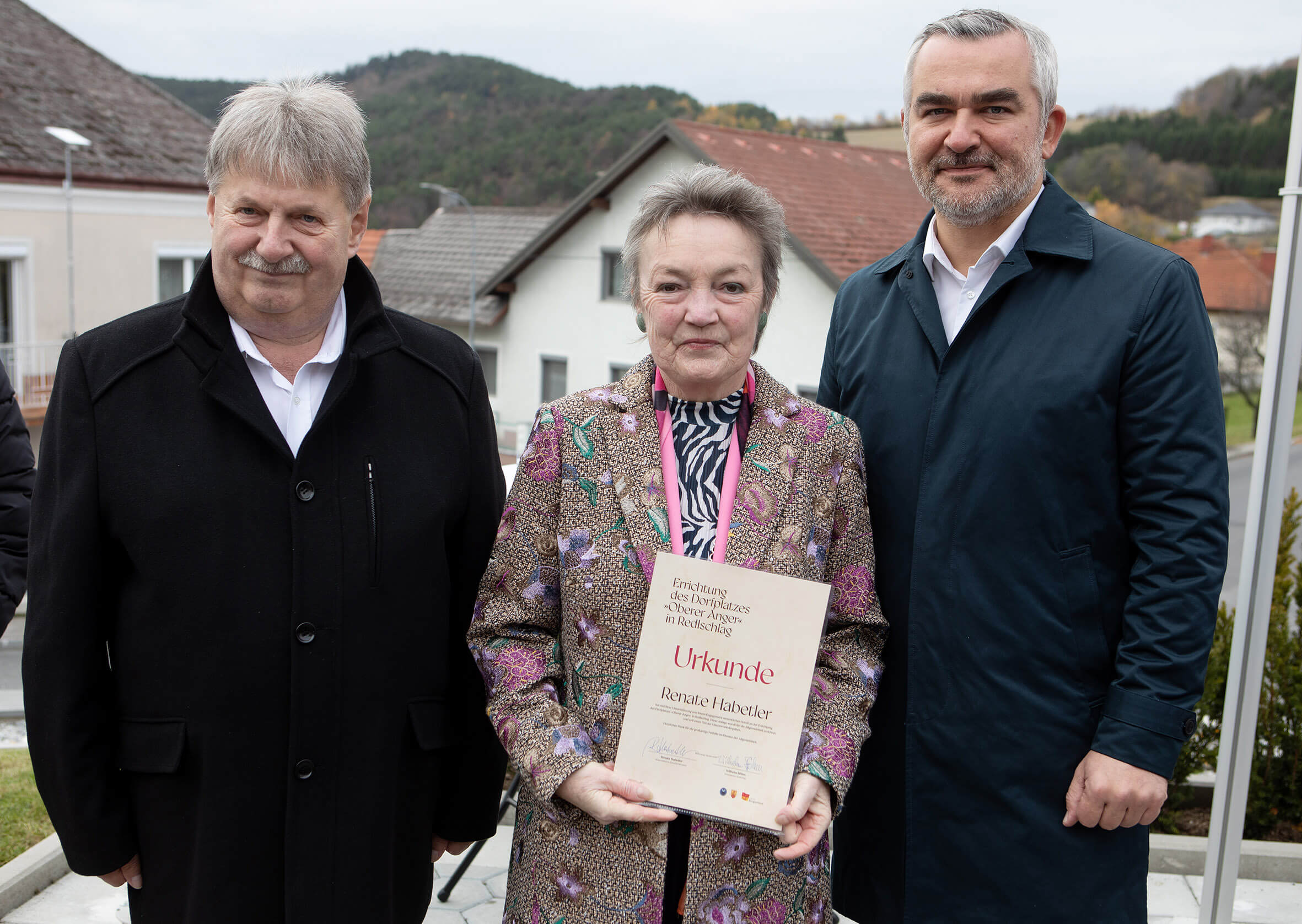 Drei Personen stehen draußen, zentrale Frau hält eine Urkunde zur Einweihung eines Dorfplatzes in Reinsberg, lächelnd.