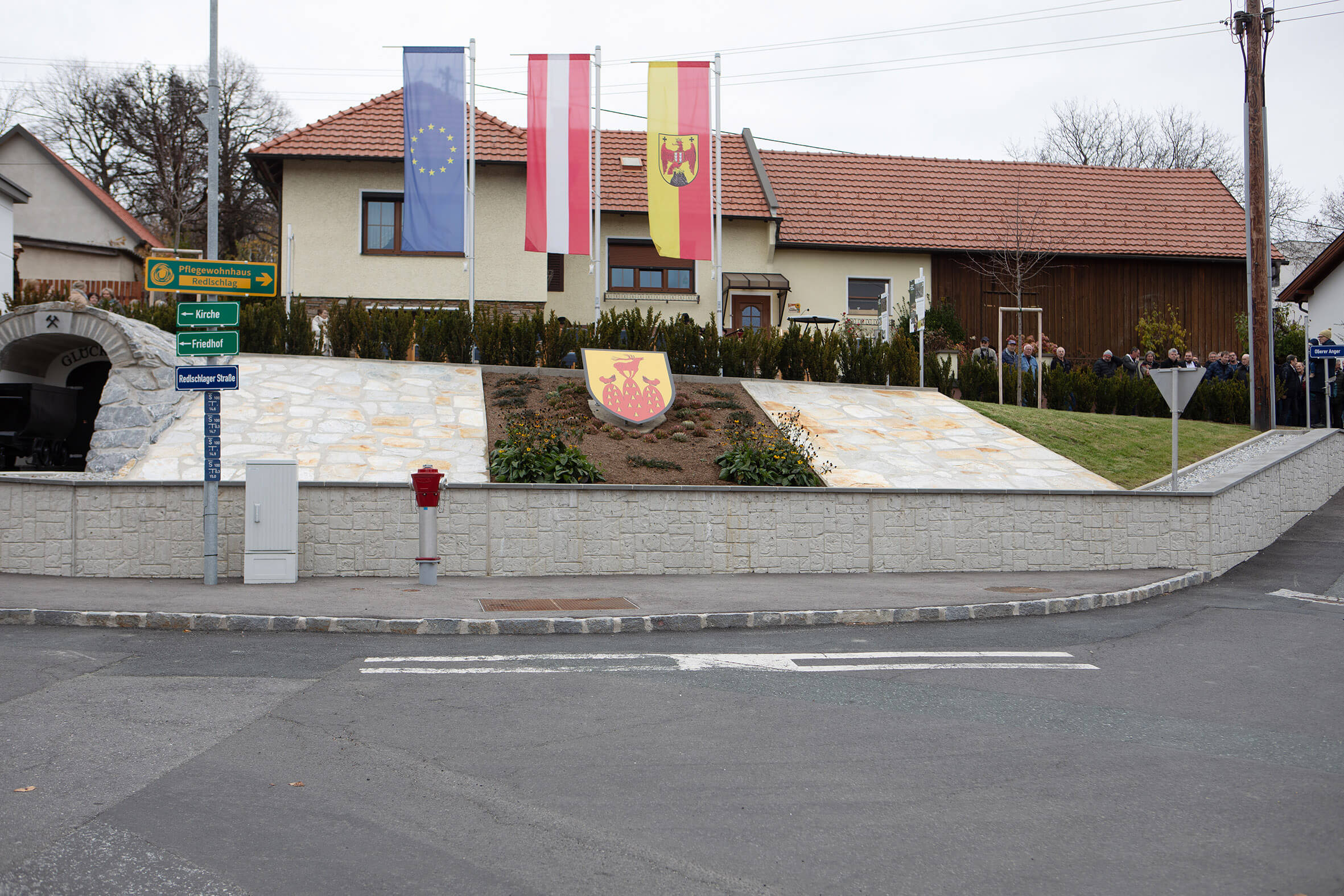 Flaggen und Wappen eines Dorfes auf einem Platz mit Straßenschildern, umgeben von Häusern.