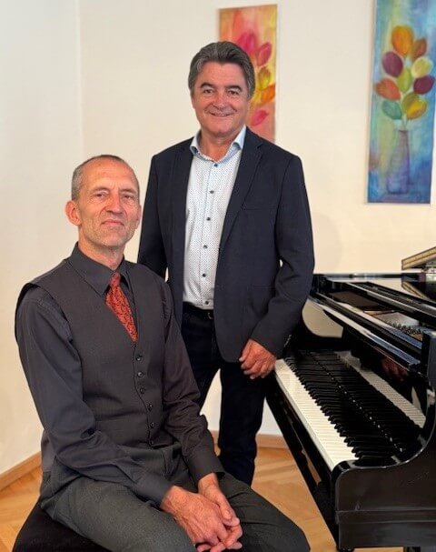 Zwei Männer posieren lächelnd vor einem Flügel in einem elegant dekorierten Raum, vermutlich für einen Artikel im Prima Magazin.