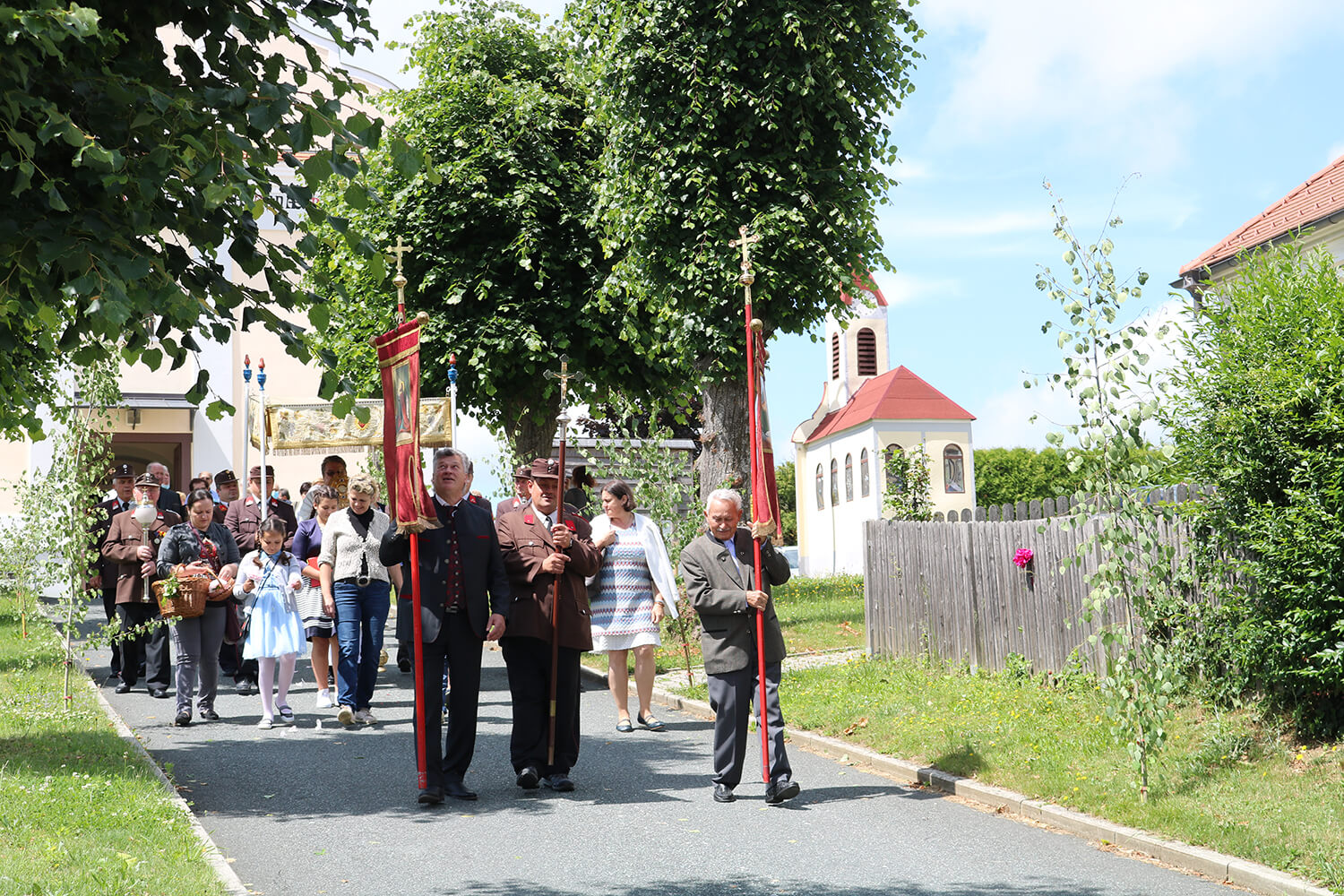 Eine traditionelle Prozession zieht an einer Dorfkirche vorbei, angeführt von Personen mit Fahnen.