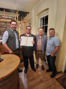 Vier Männer in traditioneller Kleidung in einem Gasthaus, einer hält eine Urkunde. Feierliche Ehrung im regionalen Kontext.