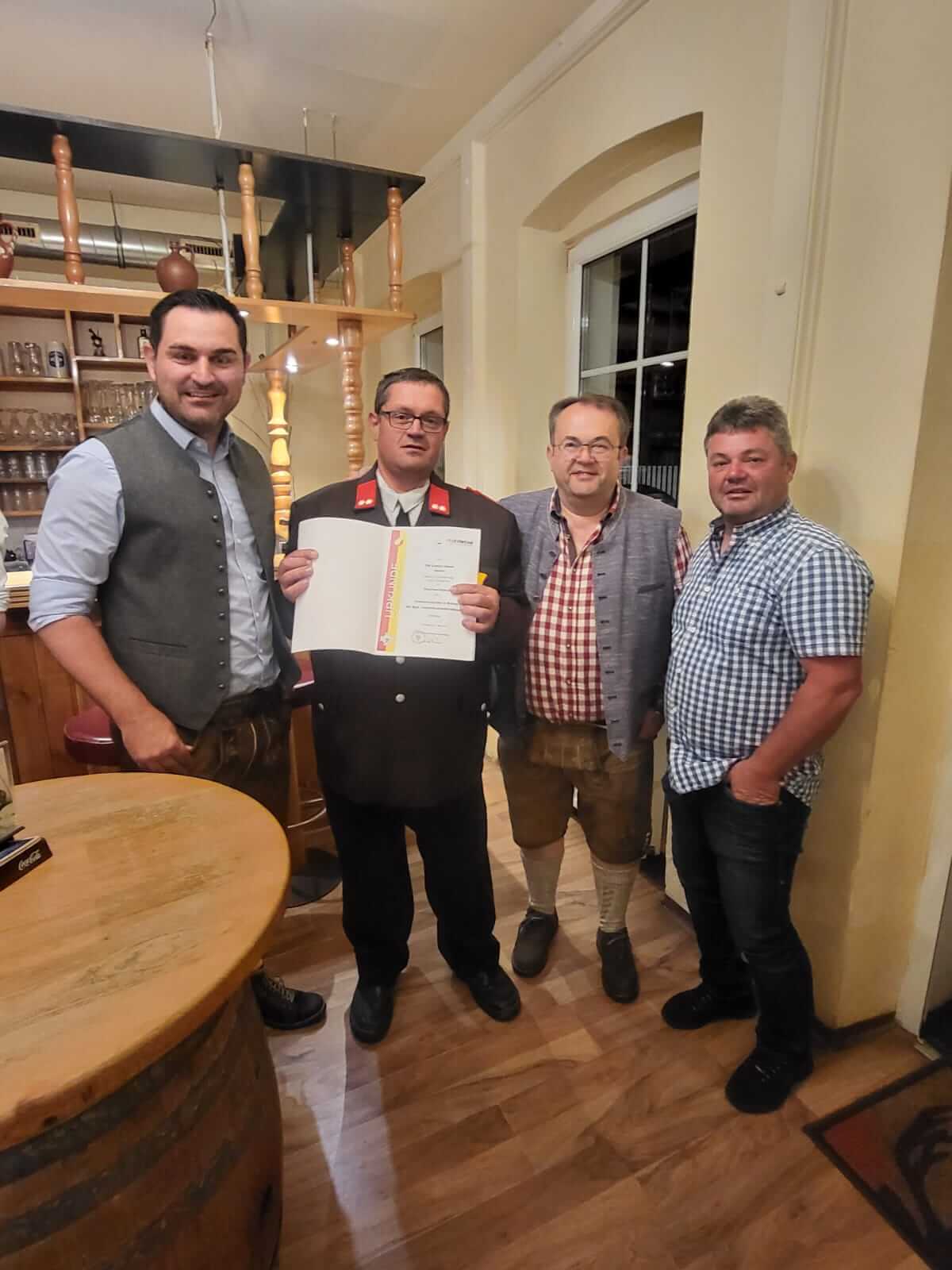 Vier Männer in traditioneller Kleidung in einem Gasthaus, einer hält eine Urkunde. Feierliche Ehrung im regionalen Kontext.