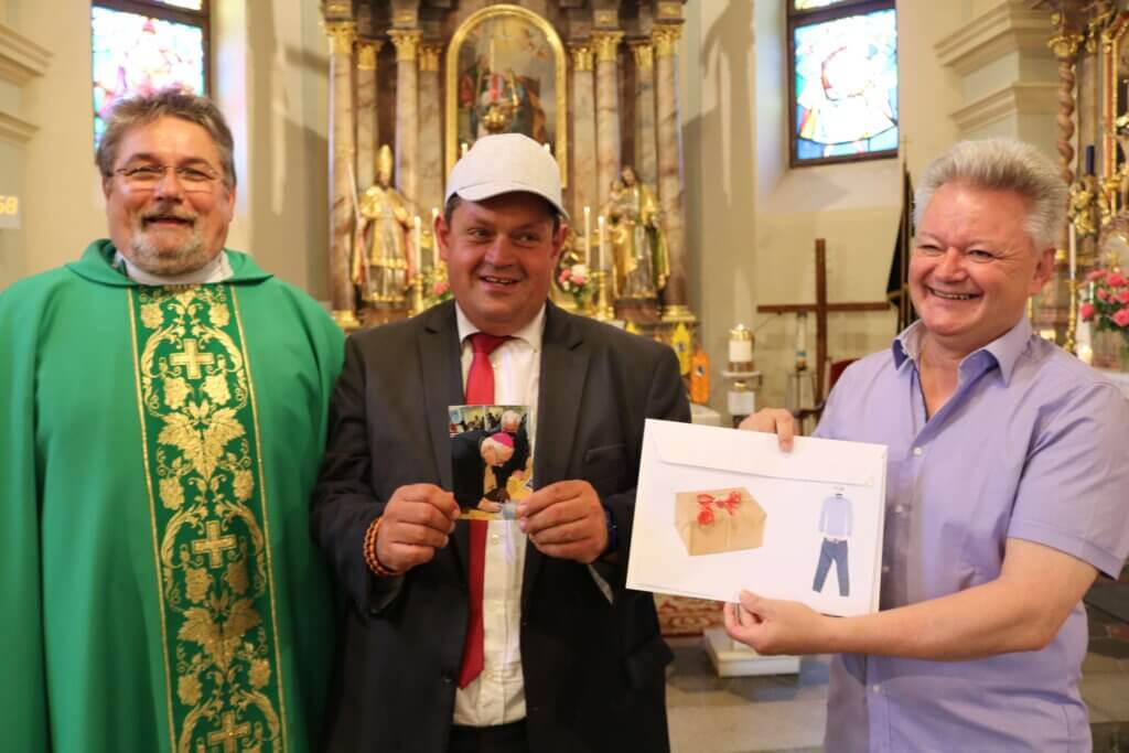 Geb. 40er (1)-c-Karl Tuider Drei Männer stehen in einer Kirche, einer in liturgischem Gewand, lächelnd ein Foto und ein Umschlag haltend.