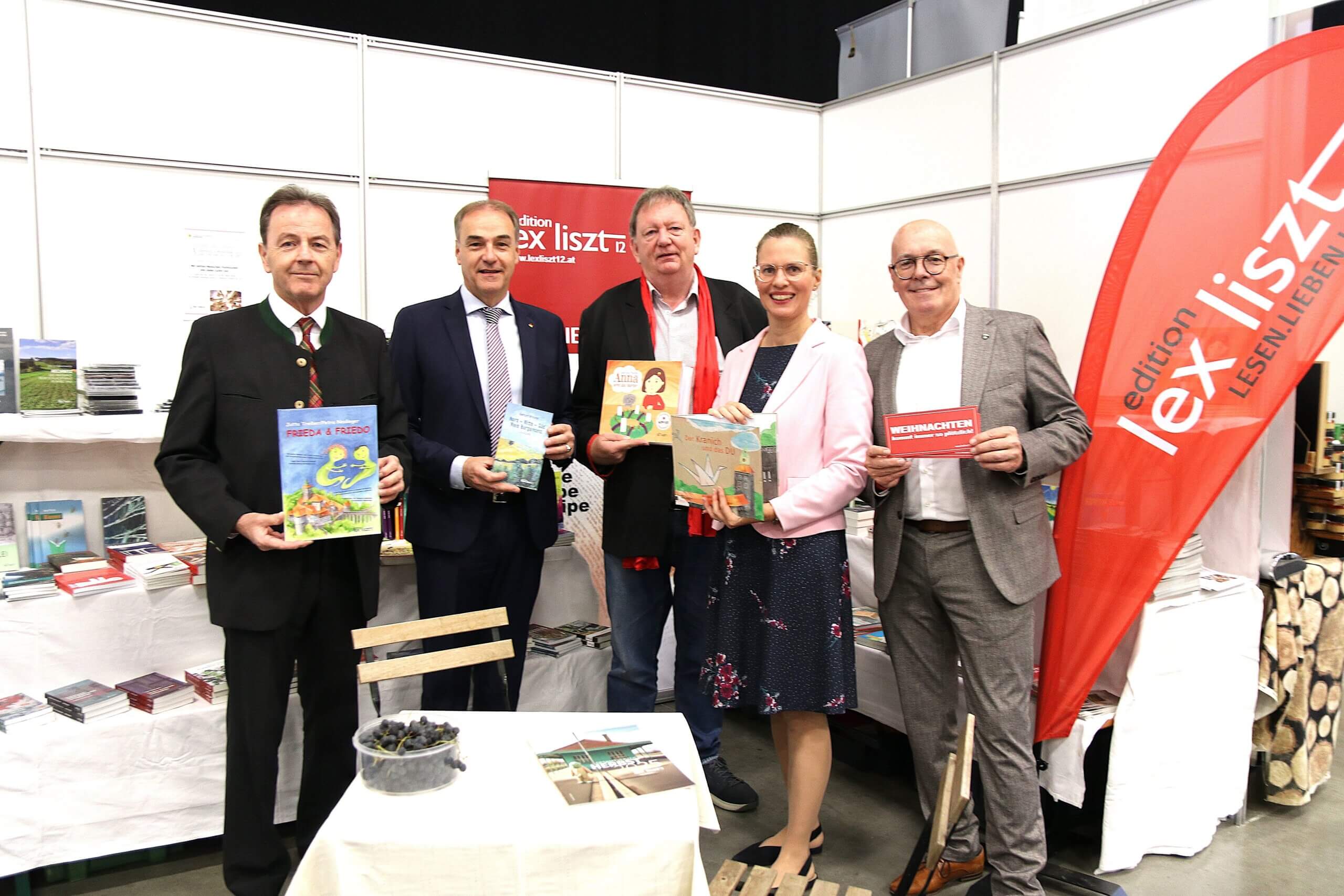 Gruppe von fünf Personen steht vor einem Buchstand, hält Bücher in den Händen und lächelt in die Kamera, bei einer Buchmesse.