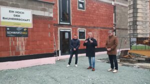 Drei Männer bei der Baustellenbesichtigung vor Rohbau mit Schild Ein Hoch der Bauherrschaft. Schwartz Bau steht im Hintergrund.