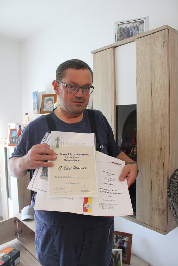 IMG_9517©Nicole Matsch Mann mit Brille hält Urkunden für langjährigen Mesnerdienst und Feuerwehrdienst vor einem Schrank, Auszeichnung gewürdigt.
