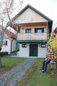 Ein Mann sitzt vor einem traditionellen Holzhaus in einer malerischen, ländlichen Umgebung.