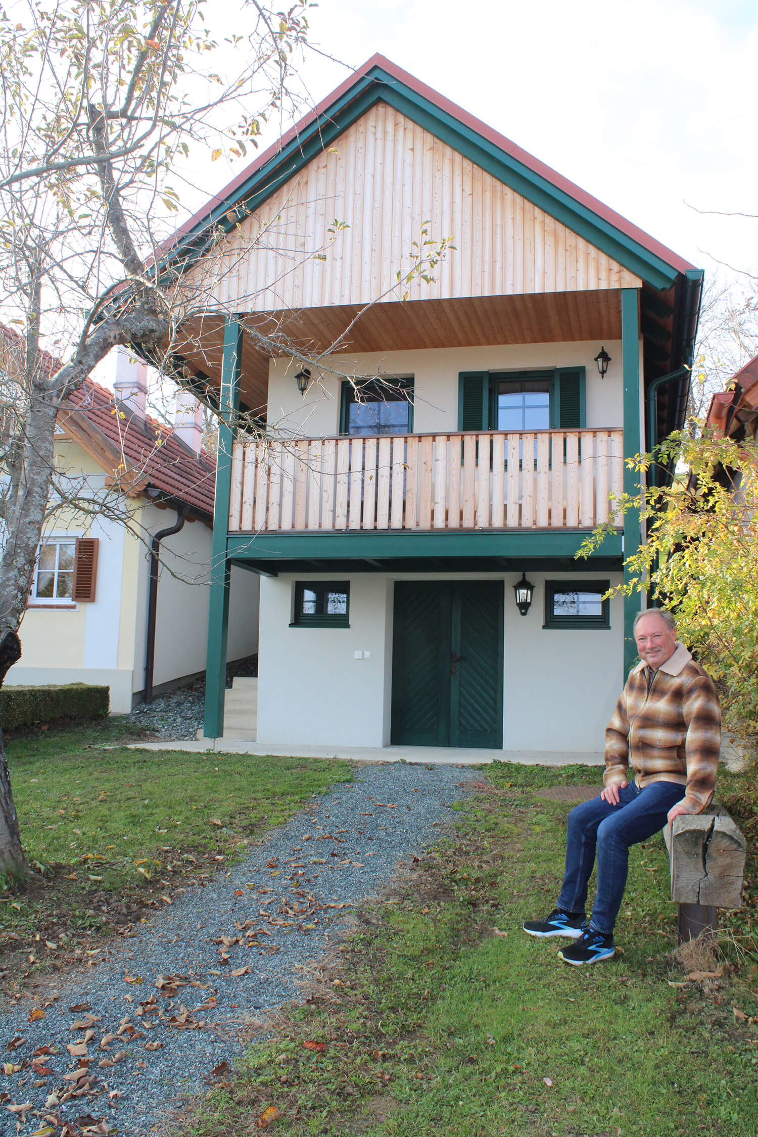 Ein Mann sitzt vor einem traditionellen Holzhaus in einer malerischen, ländlichen Umgebung.
