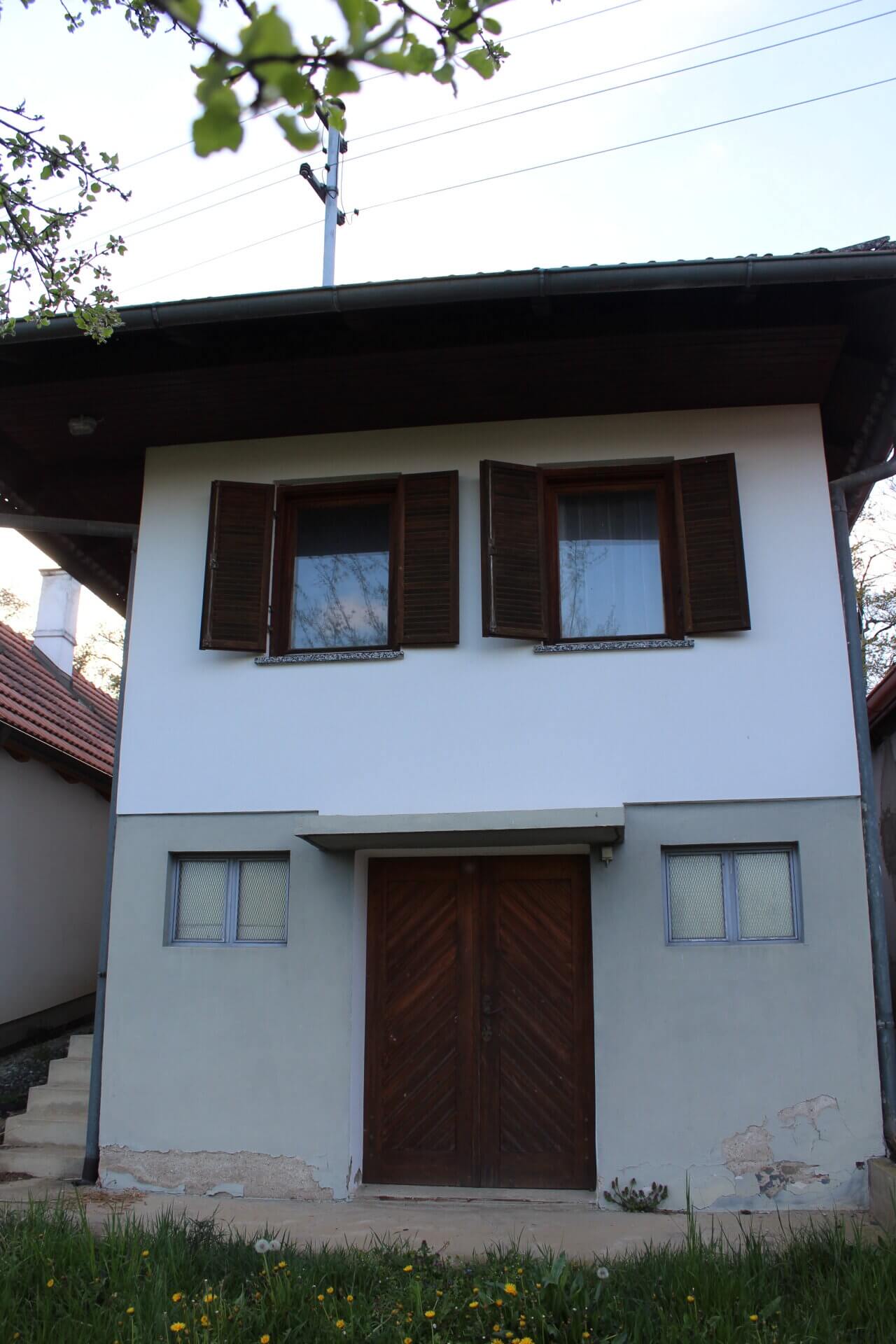 Zweistöckiges Haus mit Holztüren und Fensterläden, umgeben von frischer Natur, betont ländlichen Charme.