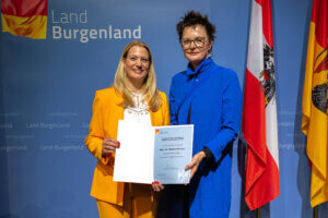 Zwei Frauen posieren stolz mit Urkunde vor einer blauen Wand des Landes Burgenland, flankiert von Landesflaggen.