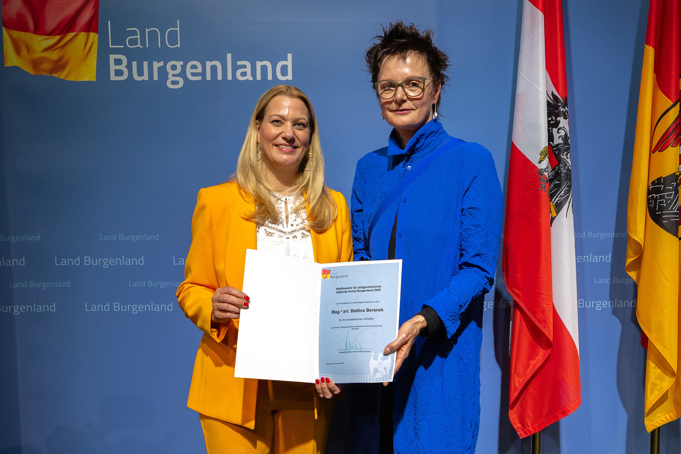 Zwei Frauen posieren stolz mit Urkunde vor einer blauen Wand des Landes Burgenland, flankiert von Landesflaggen.