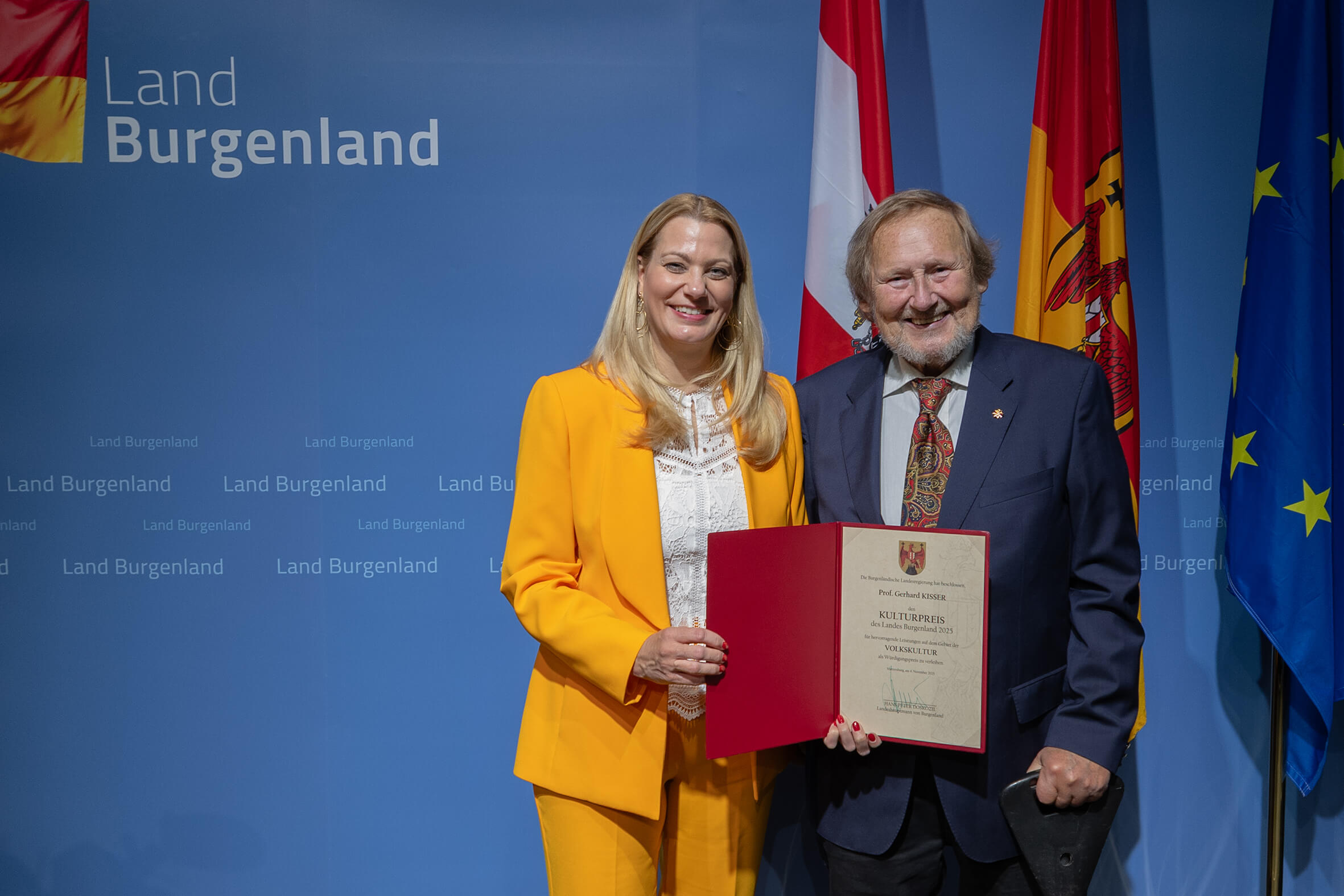 Personen mit Auszeichnung im Burgenland bei einer Preisverleihung, halten Diplom vor einer Flaggenwand; betont regionale Kultur.