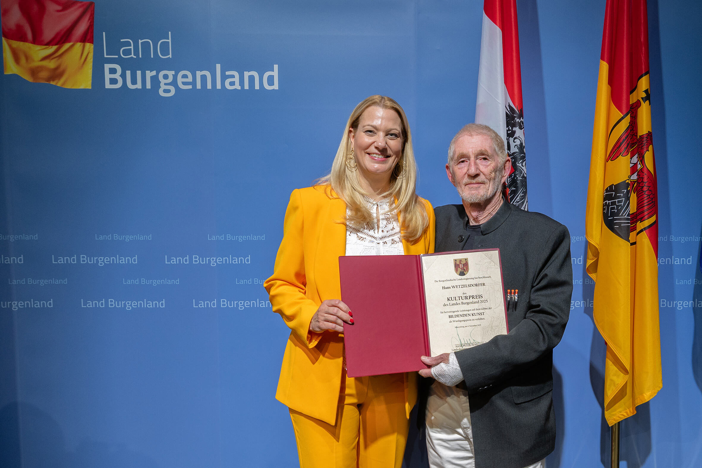 Frau in gelbem Anzug überreicht älterem Herrn eine Auszeichnung in Burgenland, Österreich, vor blauem Banner und Fahnen.