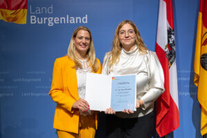 Zwei Frauen bei Preisverleihung des Landes Burgenland, halten Urkunde vor Flaggen – festlicher Moment im regionalen Kontext.