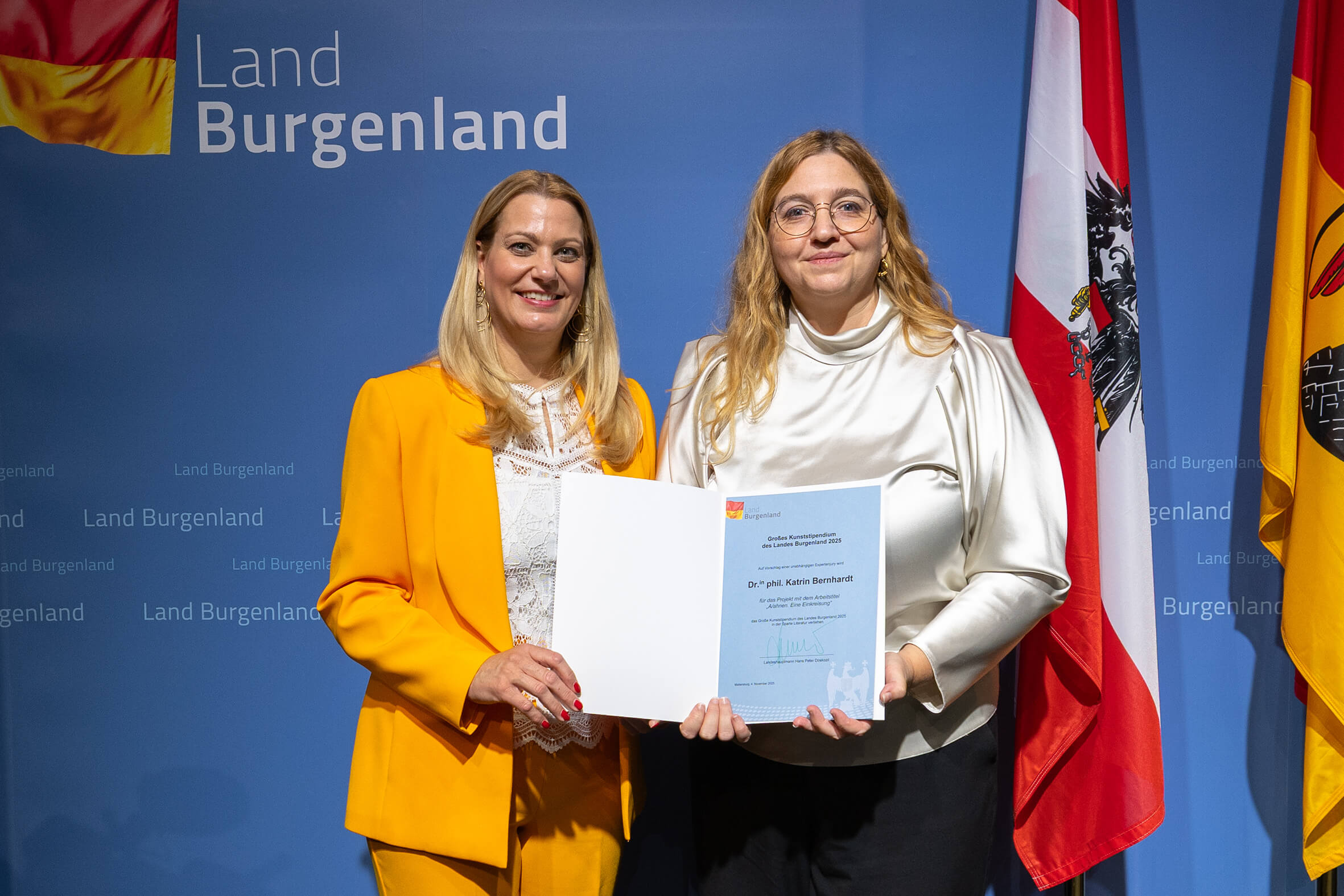 Zwei Frauen bei Preisverleihung des Landes Burgenland, halten Urkunde vor Flaggen – festlicher Moment im regionalen Kontext.