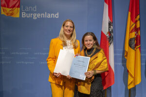 Zwei Frauen in gelben Outfits erhalten eine Urkunde vor einer Burgenland-Kulisse mit Fahnen. Feierliche Auszeichnung.
