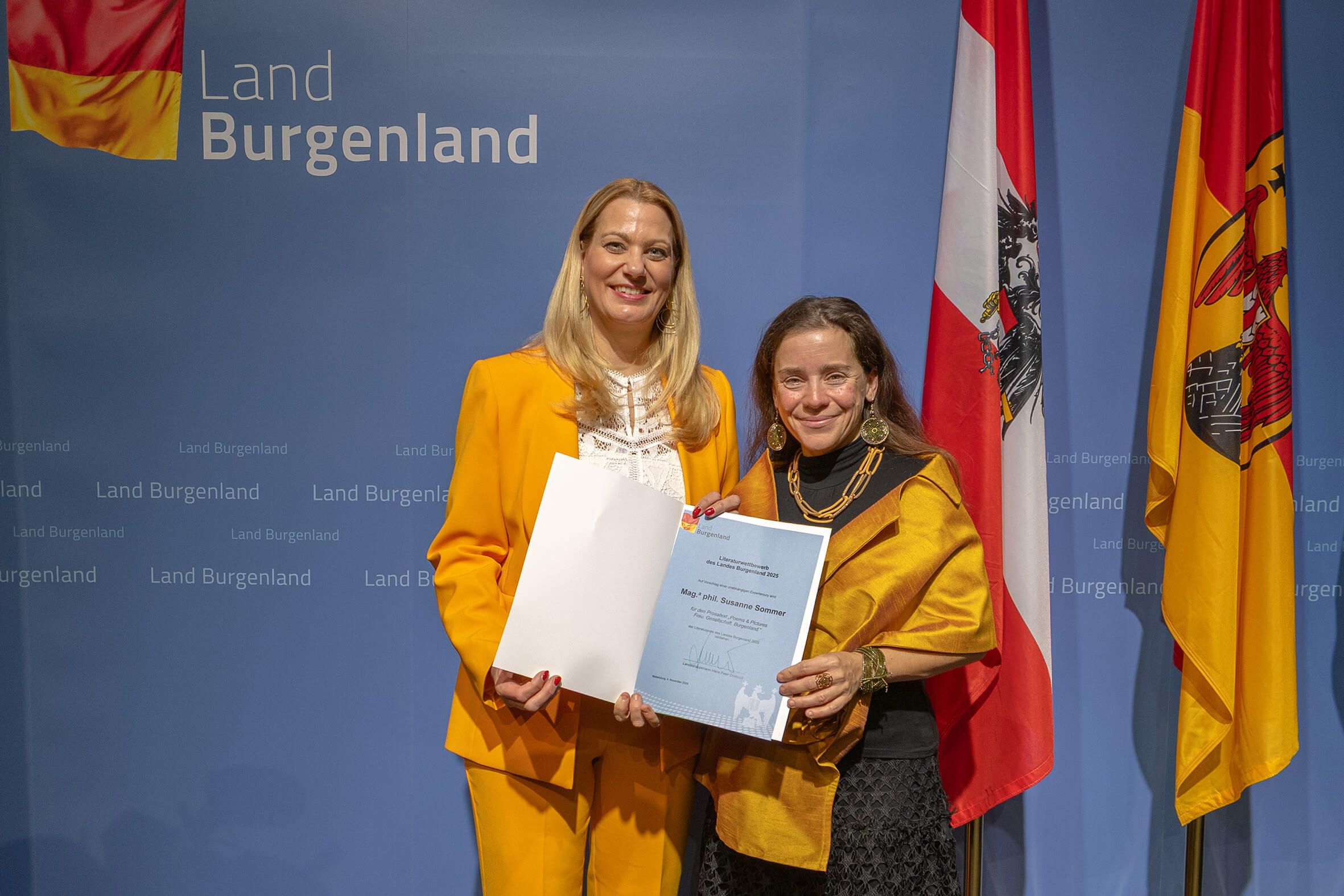Zwei Frauen in gelben Outfits erhalten eine Urkunde vor einer Burgenland-Kulisse mit Fahnen. Feierliche Auszeichnung.