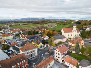 Luftaufnahme eines idyllischen österreichischen Dorfes mit Kirche und Hügeln im Hintergrund, passend für ein regionales Magazin.